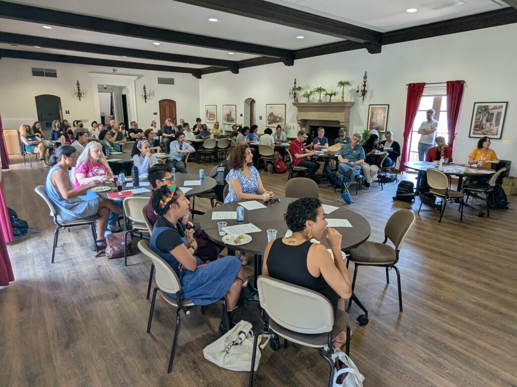 A group of some 80 people seated around tables looking at a speaker in a large hall.