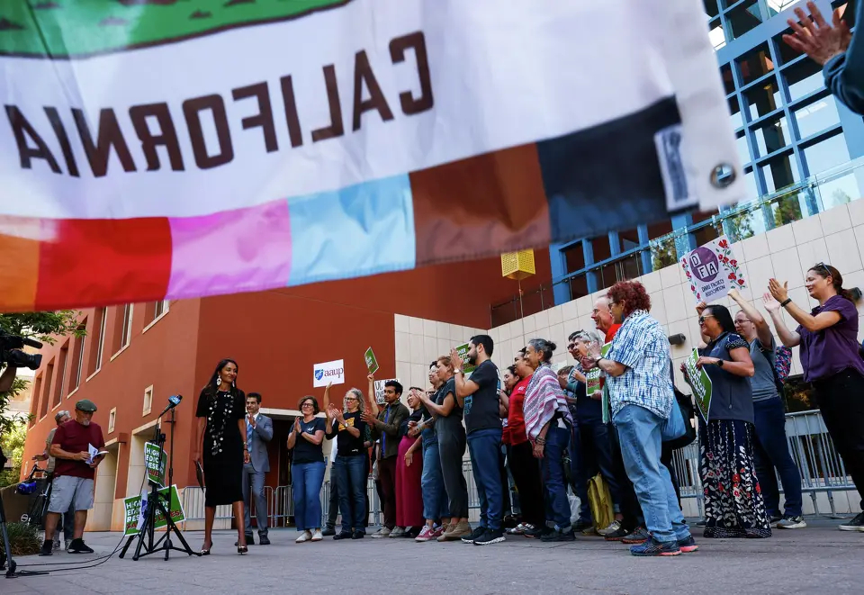 A California flag with rainbow colors stands above a group of people standing at an outside rally.